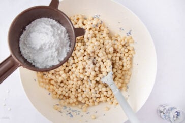 A white bowl filled with tapioca pearls and a white spatula. Above the bowl, a brown sifter holds powdered sugar, being sprinkled onto the mixture. A small clear bottle is partially visible beside the bowl.