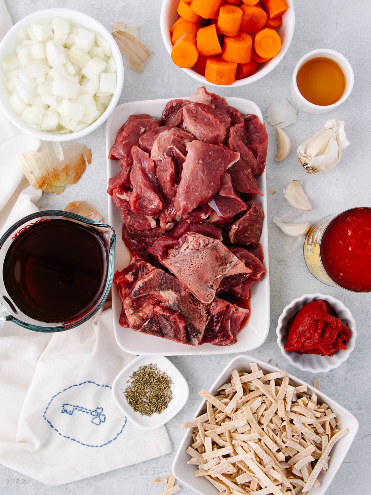 A tray of raw beef chunks is surrounded by chopped onions, sliced carrots, minced garlic, tomato paste, tomato sauce, dried herbs, red wine, broth, and sliced pasta, all arranged on a white surface.