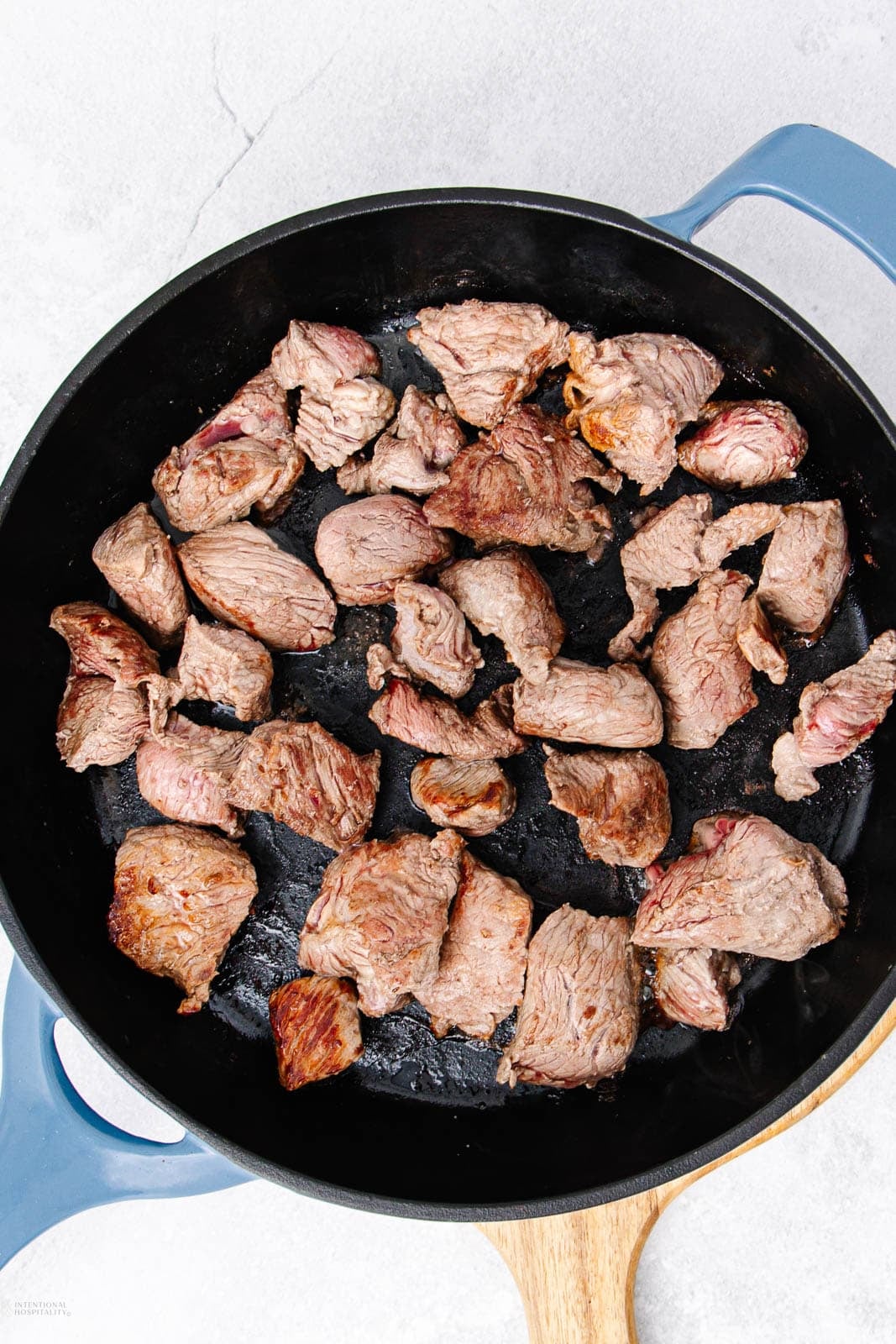 Chunks of browned beef cooking in a black skillet with blue handles, resting on a light-colored surface.
