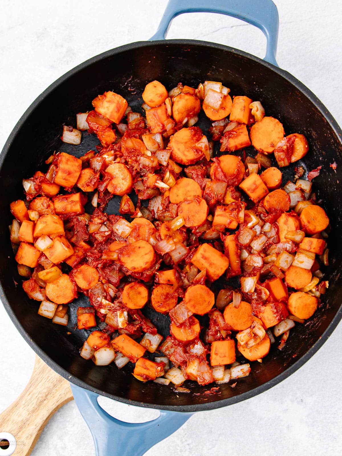 A blue-handled skillet filled with sautéed diced carrots, onions, and tomato paste sits on a white surface. The vegetables are evenly mixed and appear lightly cooked.