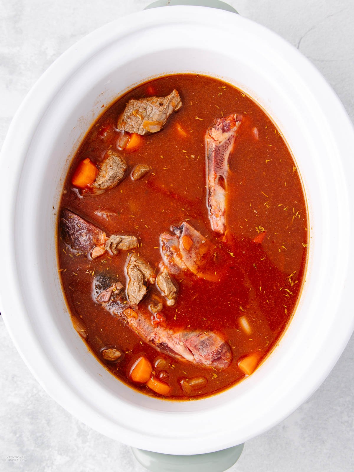 A white slow cooker filled with beef stew, featuring chunks of beef, bones, carrots, and a rich red broth. The ingredients are partially submerged and visible from the top view.