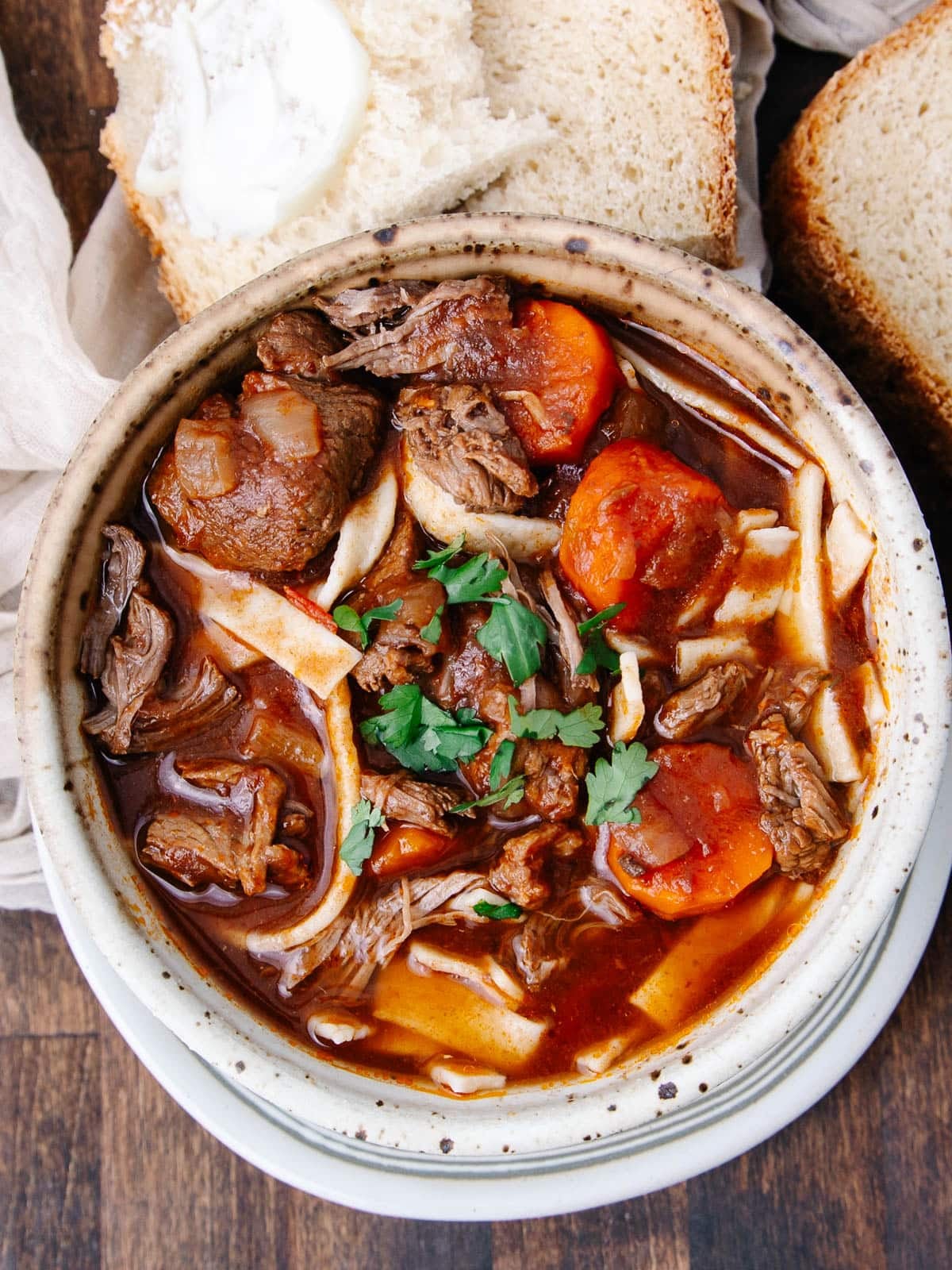 A bowl of beef noodle soup with chunks of beef, carrots, and noodles in a rich broth, garnished with fresh parsley, served with slices of buttered bread on the side.