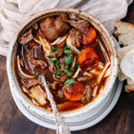 A bowl of hearty beef stew with chunks of beef, carrots, noodles, and fresh parsley, served with sliced bread on the side. A spoon is resting inside the bowl. The dish sits on a wooden table with a beige napkin nearby.