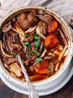 A bowl of hearty beef stew with chunks of beef, carrots, noodles, and fresh parsley, served with sliced bread on the side. A spoon is resting inside the bowl. The dish sits on a wooden table with a beige napkin nearby.