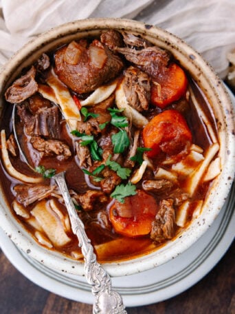 A bowl of hearty beef stew with chunks of beef, carrots, noodles, and fresh parsley, served with sliced bread on the side. A spoon is resting inside the bowl. The dish sits on a wooden table with a beige napkin nearby.