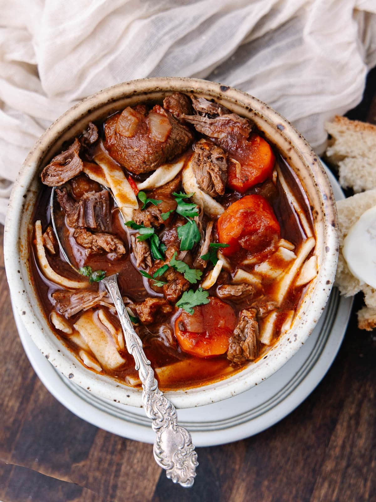 A bowl of beef stew with chunks of meat, carrots, noodles, and garnished with parsley. The stew is served with a spoon, on a wooden surface, beside a slice of bread and a light-colored cloth.