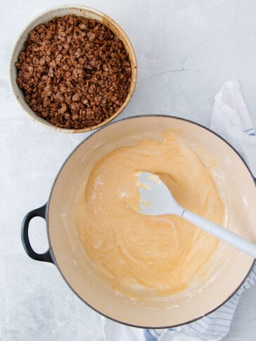 A large bowl with creamy batter and a spatula sits next to a smaller bowl filled with chocolate crispy cereal on a light countertop. A white kitchen towel with blue stripes is nearby.