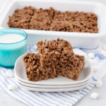 Three chocolate crispy rice treats stacked on plates next to a glass of milk, with more treats in a white baking dish behind. Mini marshmallows are scattered on a striped cloth underneath.