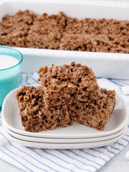 Three chocolate crispy rice treats stacked on plates next to a glass of milk, with more treats in a white baking dish behind. Mini marshmallows are scattered on a striped cloth underneath.