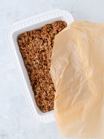 A white rectangular baking dish lined with brown parchment paper, containing a chocolate granola mixture pressed evenly inside. The parchment paper covers about half of the granola.