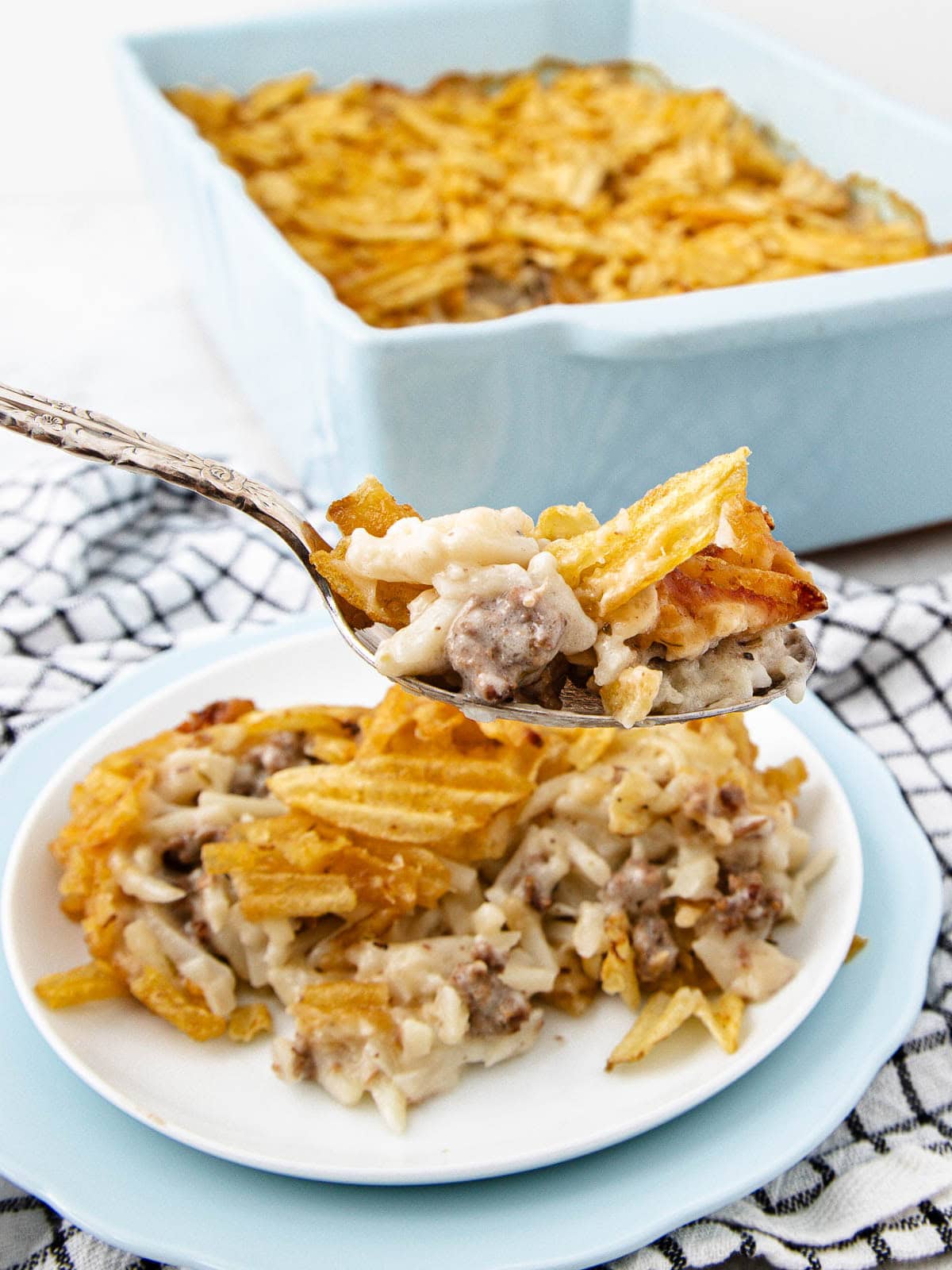 A serving spoon holds up cheesy ground beef and noodle casserole topped with crispy potato chips, with a plate of the casserole beneath and a baking dish in the background.