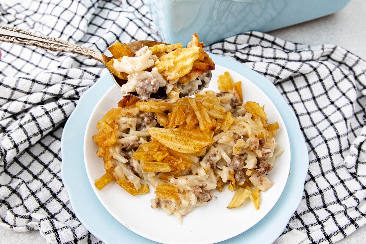 A plate of cheesy casserole with ground beef, topped with crispy potato chips, sits on a light blue plate. A serving spoon lifts a portion above the plate. A checkered cloth and blue dish are in the background.