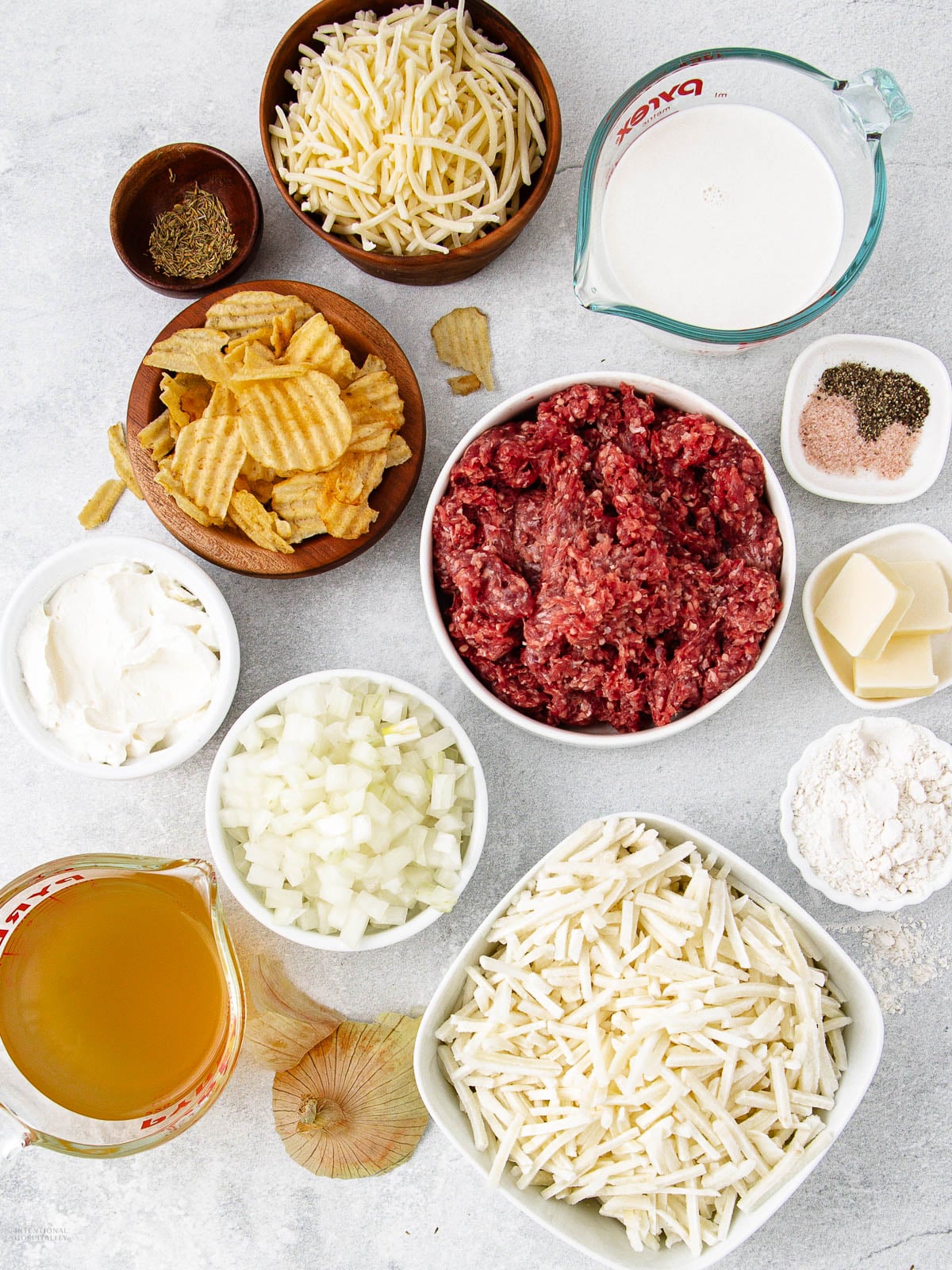 Ingredients for a casserole recipe arranged on a light surface, including shredded cheese, ground beef, chopped onions, hash browns, potato chips, broth, cream, spices, flour, butter, and cream cheese.