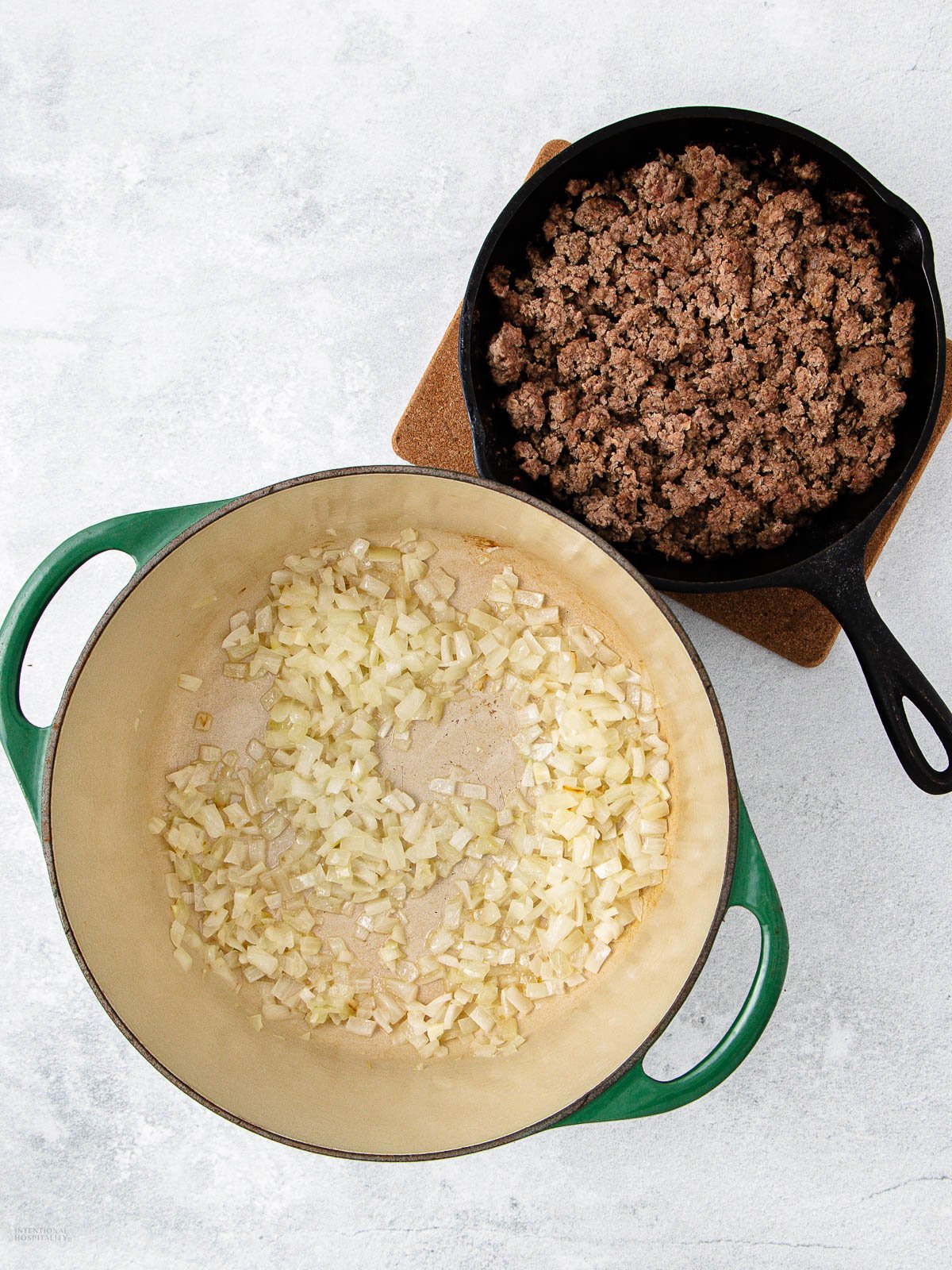A green Dutch oven with sautéed chopped onions sits next to a black skillet filled with cooked ground beef on a light countertop.