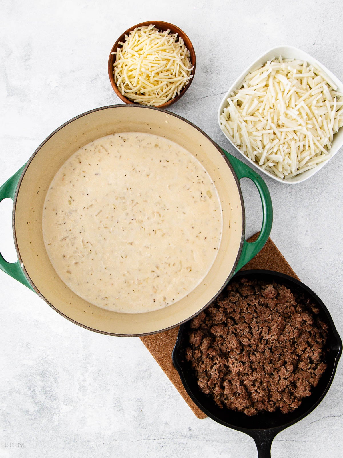 A pot filled with creamy soup sits on a trivet, surrounded by a skillet of cooked ground beef, a bowl of shredded cheese, and a bowl of frozen hash browns on a white surface.