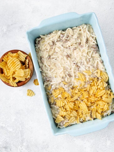 A casserole dish filled with a creamy mixture topped halfway with crumbled ridged potato chips, with a small bowl of additional potato chips beside it on a light-colored surface.