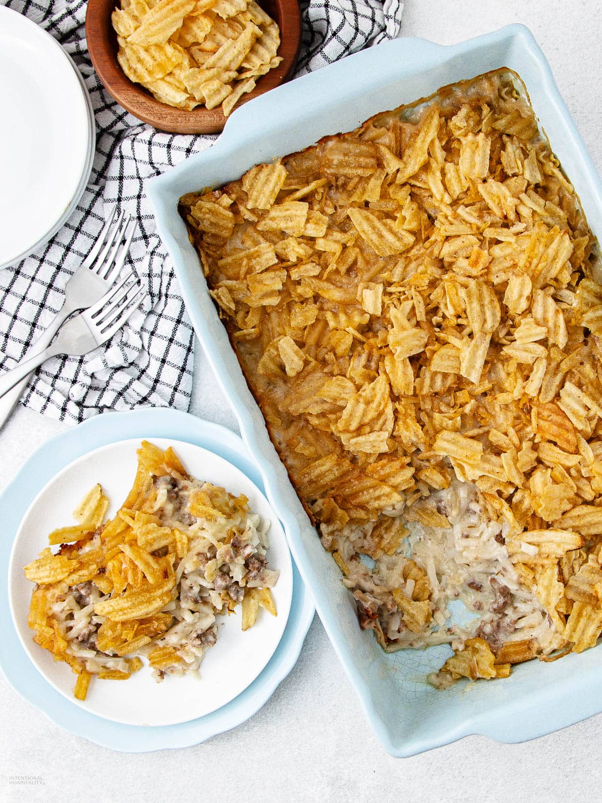 A baking dish filled with a casserole topped with crinkled potato chips, with one serving on a white plate nearby. A small bowl of chips, a checkered cloth, and utensils are also visible.