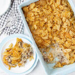 A rectangular casserole dish filled with a baked hash brown casserole topped with crispy potato chips, with a portion served on a white plate nearby. A checkered cloth and forks are beside the dish.