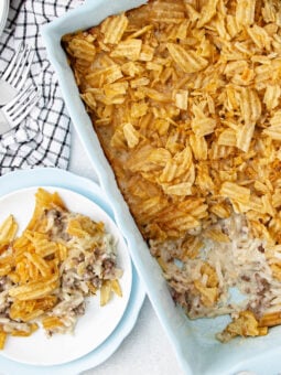 A rectangular casserole dish filled with a baked hash brown casserole topped with crispy potato chips, with a portion served on a white plate nearby. A checkered cloth and forks are beside the dish.
