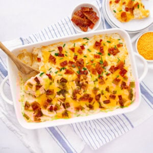 A white baking dish filled with loaded baked potato casserole—cheesy mashed potatoes topped with chopped bacon and chives—sits ready, as a wooden spoon scoops some out. Nearby, a bowl of bacon and shredded cheese rests on a striped cloth.