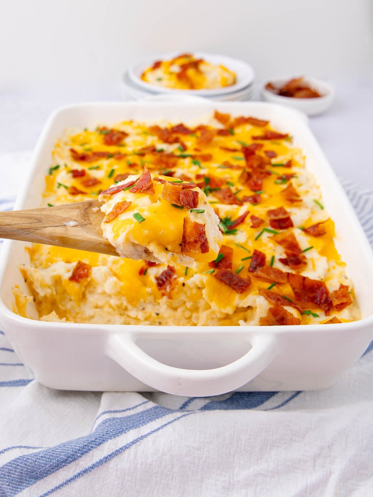 A wooden spoon lifts a serving of cheesy mashed potato casserole topped with melted cheese, chopped bacon, and chives from a white baking dish. The dish sits on a striped cloth with small bowls in the background.