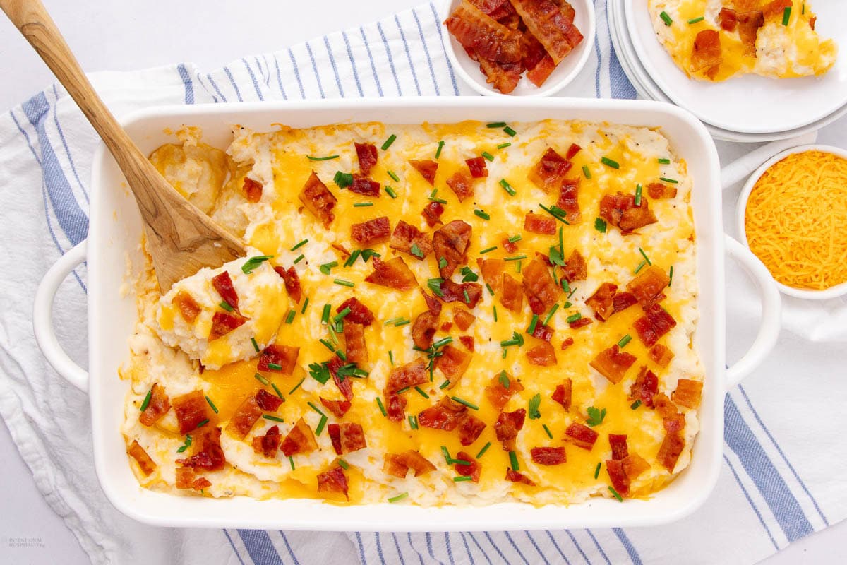 A casserole dish of loaded mashed potatoes topped with melted cheese, crispy bacon bits, and chopped chives. A wooden spoon serves a portion. Bowls of shredded cheese and extra servings are nearby on a striped cloth.