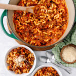 A pot of macaroni with ground beef and tomato sauce sits on a table next to two bowls of the dish, grated cheese, two spoons, and a green checkered napkin. One bowl is topped with shredded cheese.