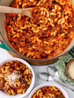 A pot of macaroni with ground beef and tomato sauce sits on a table next to two bowls of the dish, grated cheese, two spoons, and a green checkered napkin. One bowl is topped with shredded cheese.