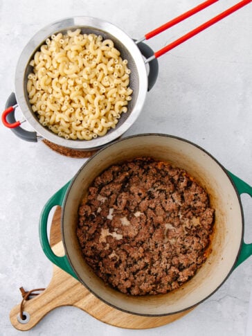 A colander with cooked elbow macaroni sits above a green pot filled with browned ground beef, both placed on a light-colored countertop with a wooden cutting board underneath the pot.