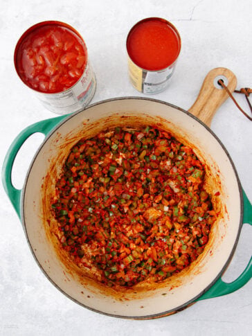 A green Dutch oven filled with sautéed chopped vegetables sits on a white surface beside two open cans of tomatoes—one with diced tomatoes and the other with tomato puree. A wooden spoon rests nearby.
