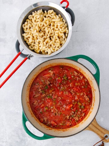 A green pot of tomato-based meat sauce with peppers sits beside a colander of cooked macaroni pasta on a white surface.