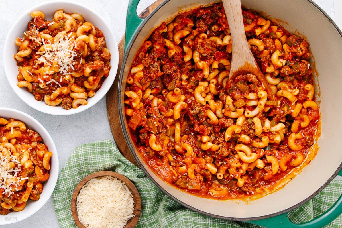 A pot of elbow macaroni with meat sauce sits on a green cloth, with a wooden spoon inside. Two bowls of the pasta are nearby, each topped with grated cheese, and a small bowl of shredded cheese is on the side.