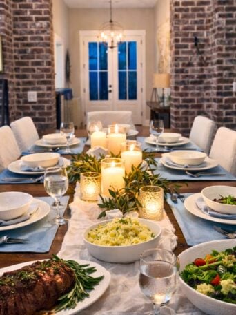 A warmly lit dining table set for dinner with white plates, bowls of salad, mashed potatoes, a platter of meat, water glasses, candles, and greenery, in a cozy room with brick walls and double doors in the background.