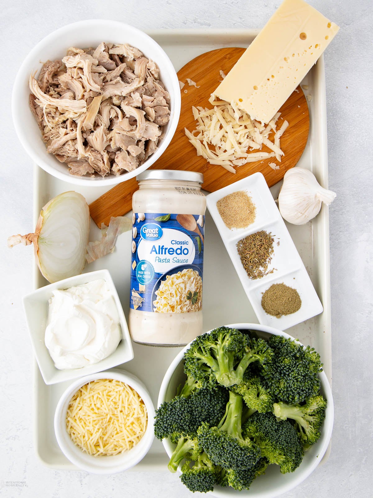 A tray with bowls of shredded chicken, broccoli florets, shredded cheese, cream cheese, grated parmesan, a jar of Alfredo sauce, half an onion, garlic, spices, and a block of cheese arranged neatly.