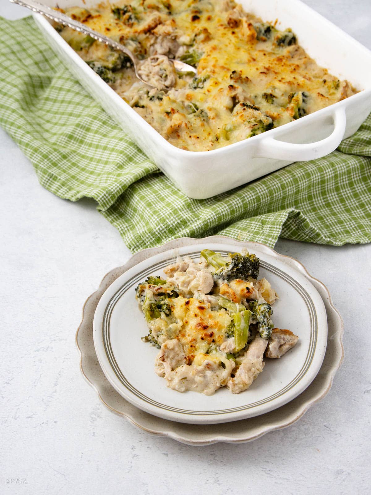 A plate of cheesy chicken and broccoli casserole sits in front of a white baking dish filled with more casserole, all placed on a green checkered cloth.