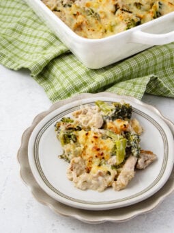 A serving of cheesy chicken and broccoli casserole on a white plate, with the casserole dish and a green checkered cloth in the background. The dish appears creamy and baked with a golden top.
