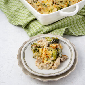 A serving of cheesy chicken and broccoli casserole on a white plate, with the casserole dish and a green checkered cloth in the background. The dish appears creamy and baked with a golden top.