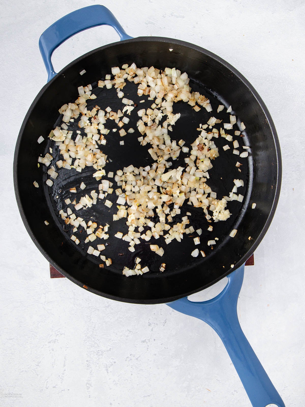 Diced onions being sautéed in a large black skillet with a blue handle, set on a light gray surface. The onions are lightly browned and spread across the pan.