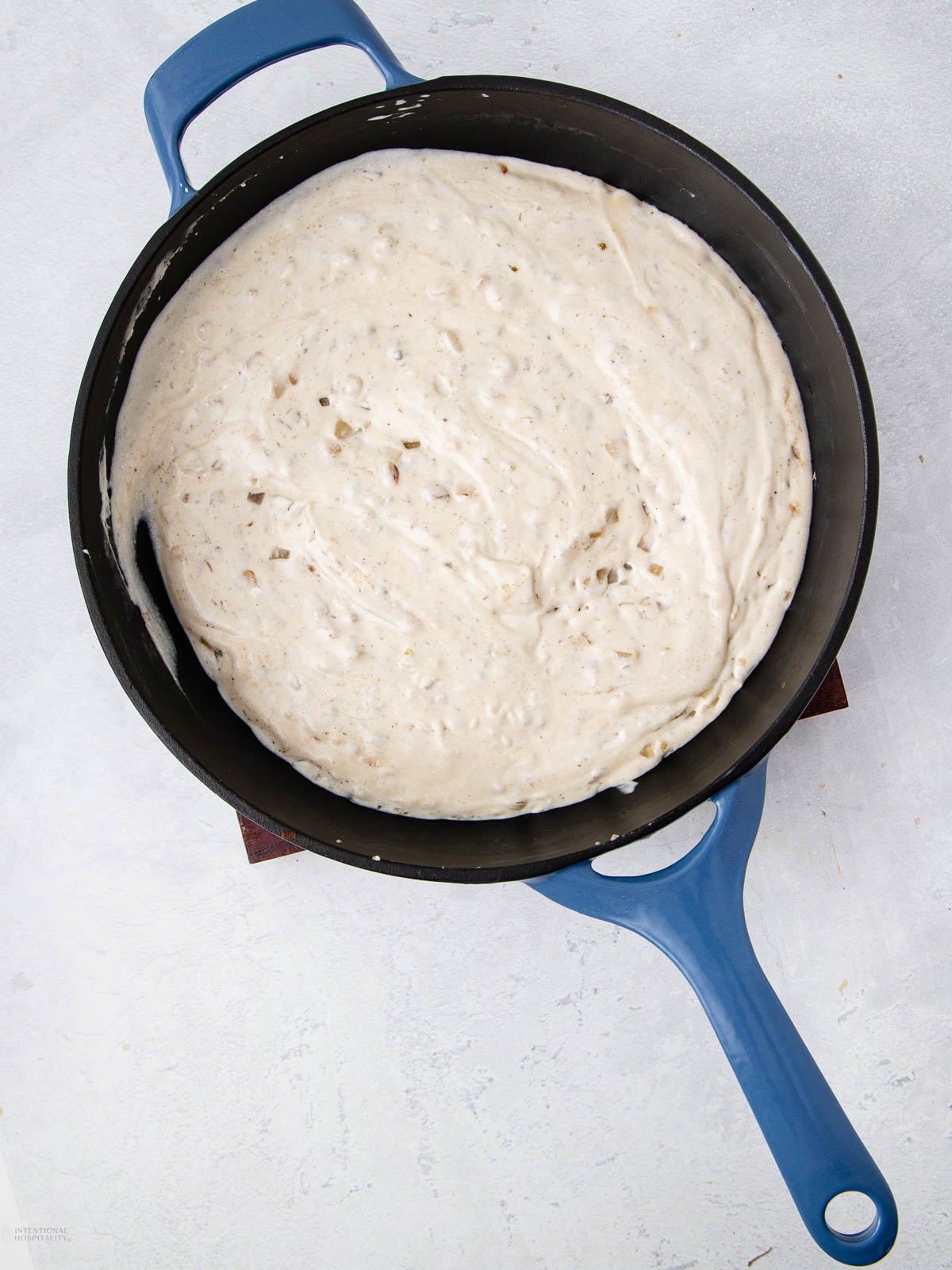 A blue-handled cast iron pan filled with bubbly, risen bread dough sits on a light-colored surface.
