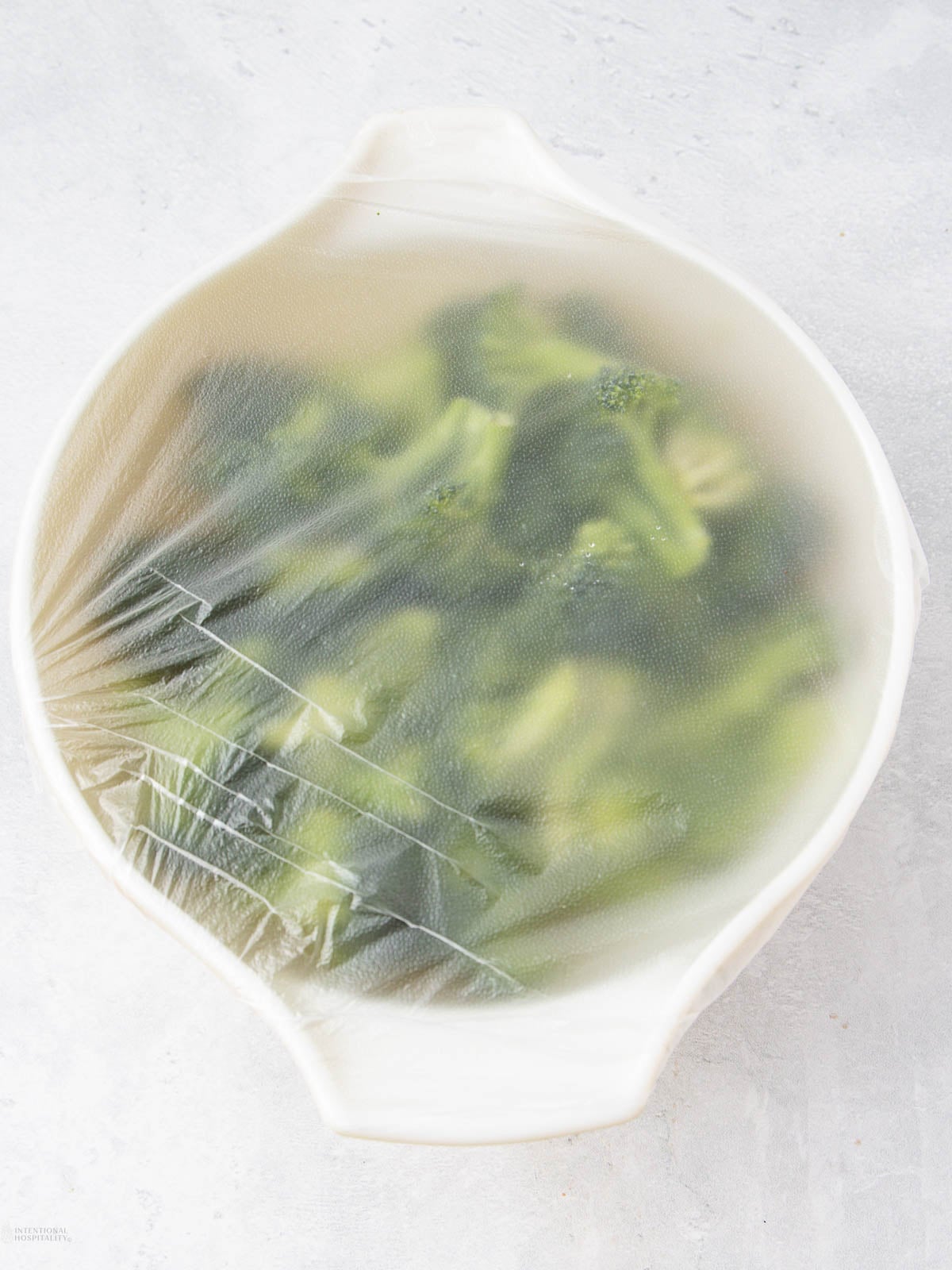 A white bowl filled with broccoli florets is covered tightly with plastic wrap, set on a light-colored surface.