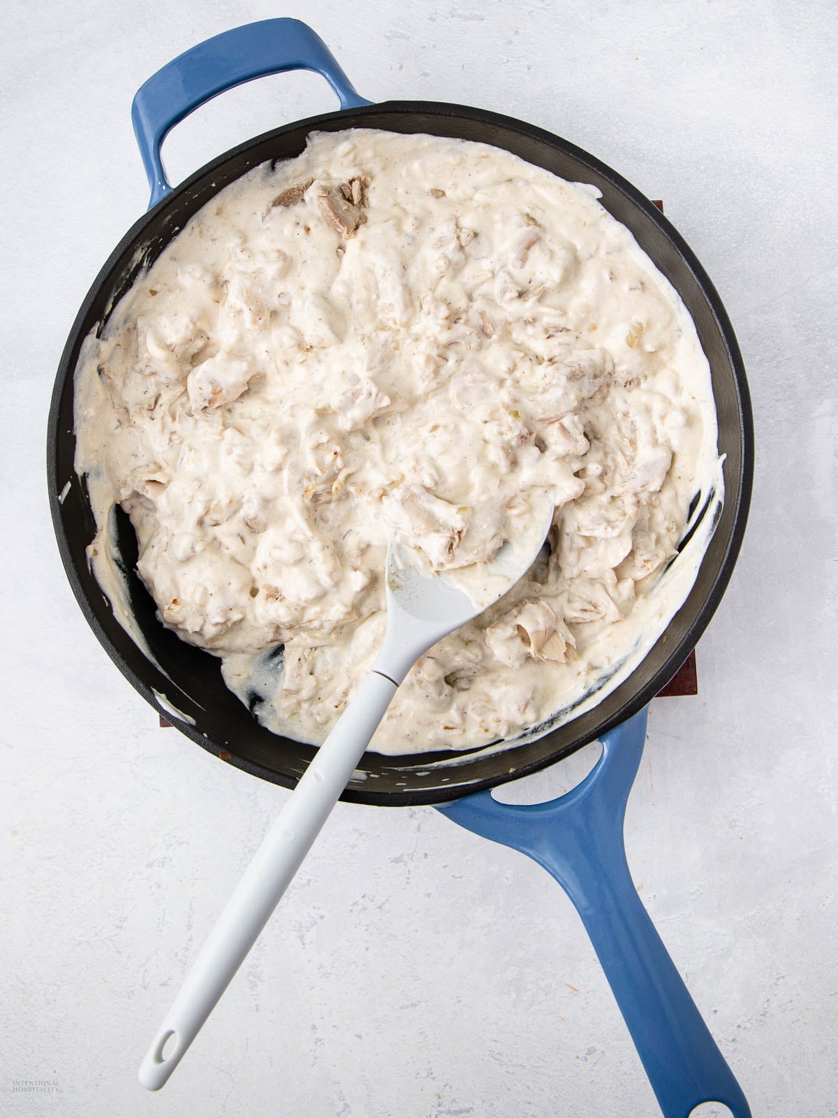 A blue-handled skillet filled with creamy shredded chicken mixture, being stirred with a white spatula, sits on a light-colored surface.