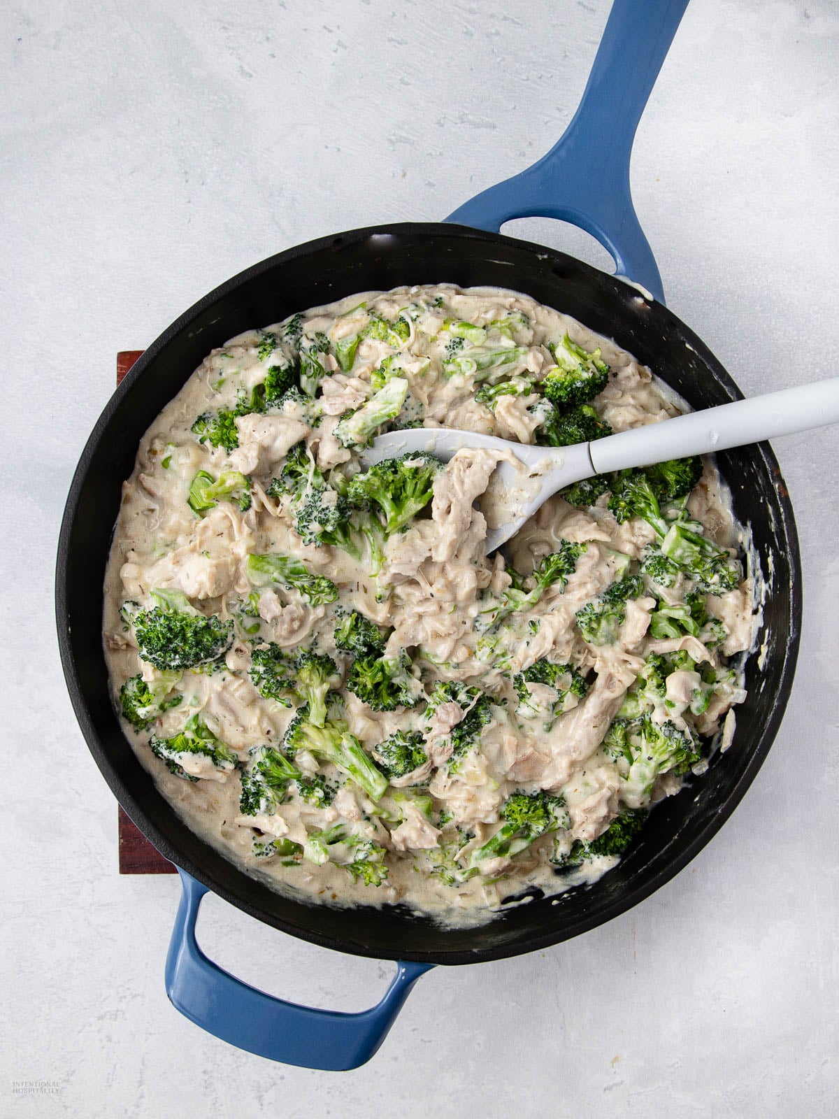 A blue skillet filled with a creamy chicken and broccoli mixture, being stirred with a white spatula, on a light gray surface.
