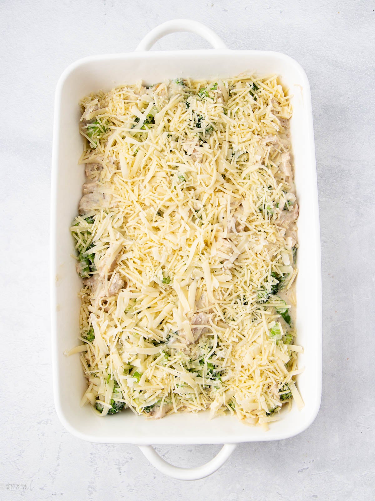 A white rectangular baking dish filled with an uncooked casserole topped with shredded cheese, visible pieces of green vegetables and creamy sauce underneath. The dish sits on a light gray surface.