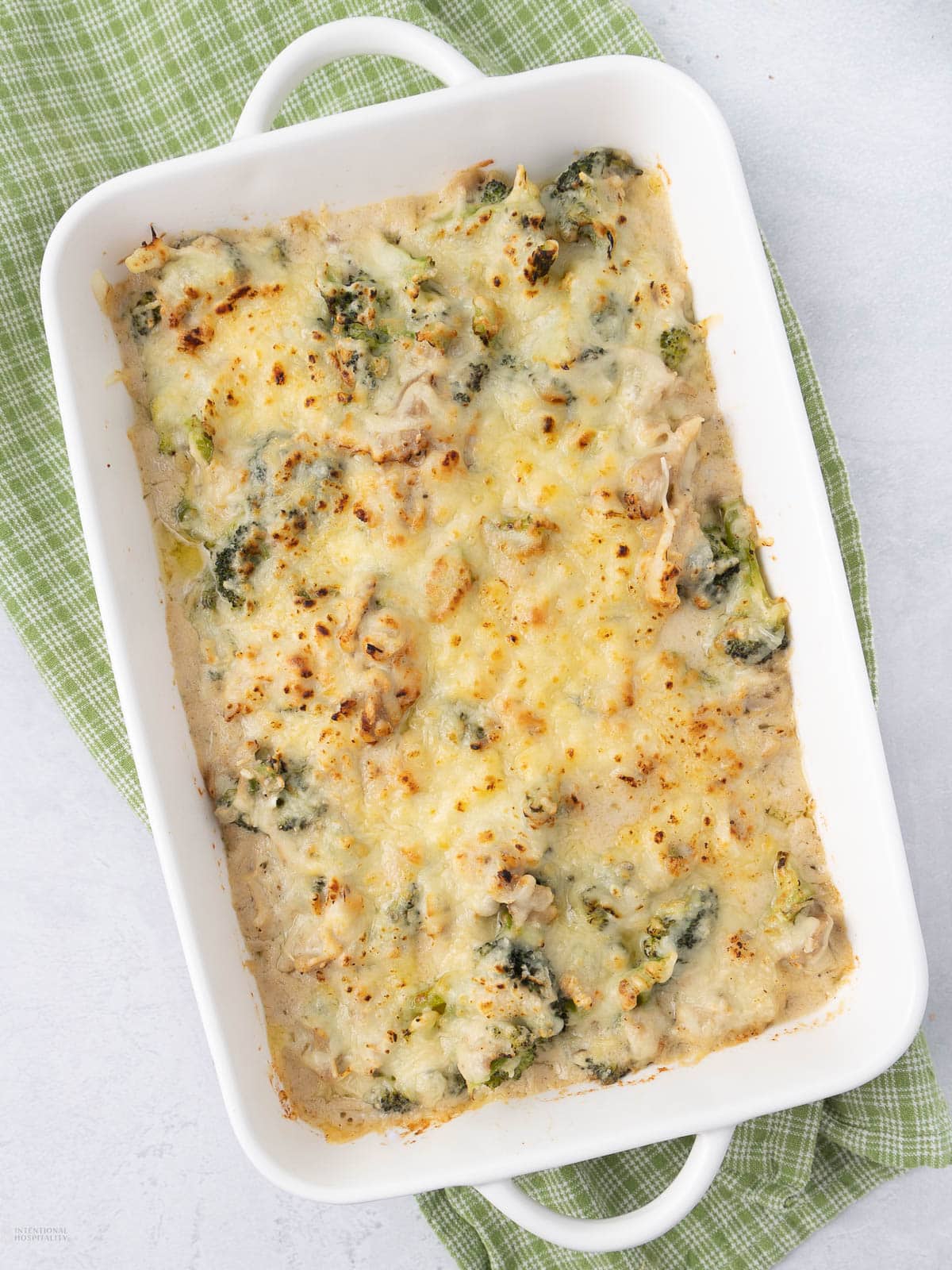 A white rectangular baking dish filled with cheesy broccoli casserole sits on a green checkered cloth, viewed from above on a light-colored surface.