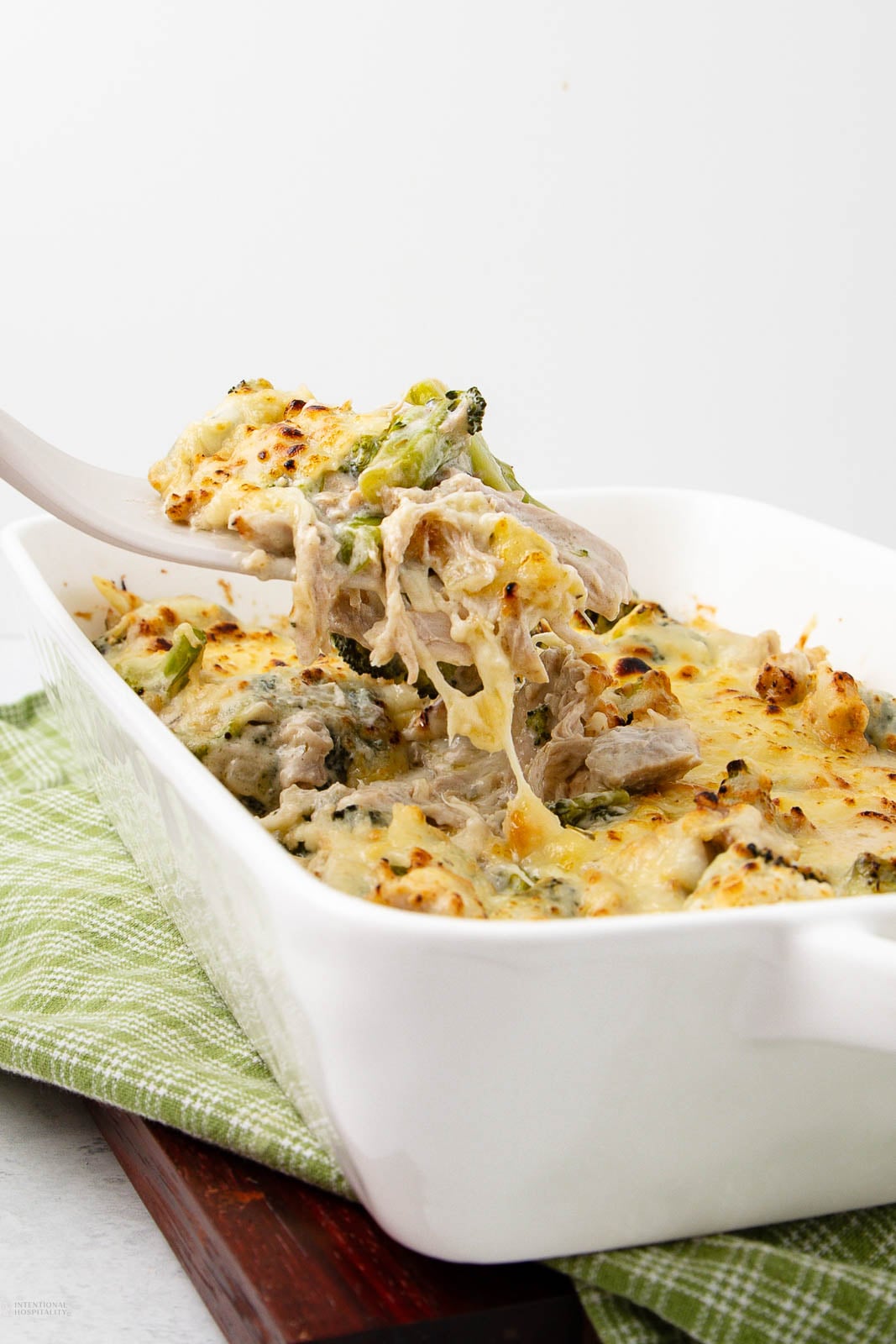 A serving spoon lifts a cheesy, baked chicken and broccoli casserole from a white dish, showing melted cheese and shredded chicken. The dish rests on a green plaid cloth and wooden board.