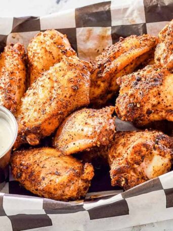 A basket of seasoned chicken wings on checkered paper, served with a cup of creamy dipping sauce on the side.