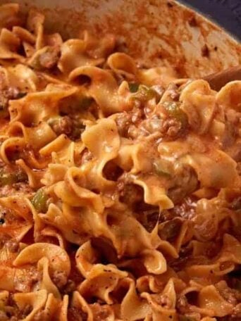 A wooden spoon scoops up cheesy beef and noodle casserole from a pot. The dish features curly egg noodles, ground beef, green bell peppers, and a tomato-based sauce, topped with melted cheese.