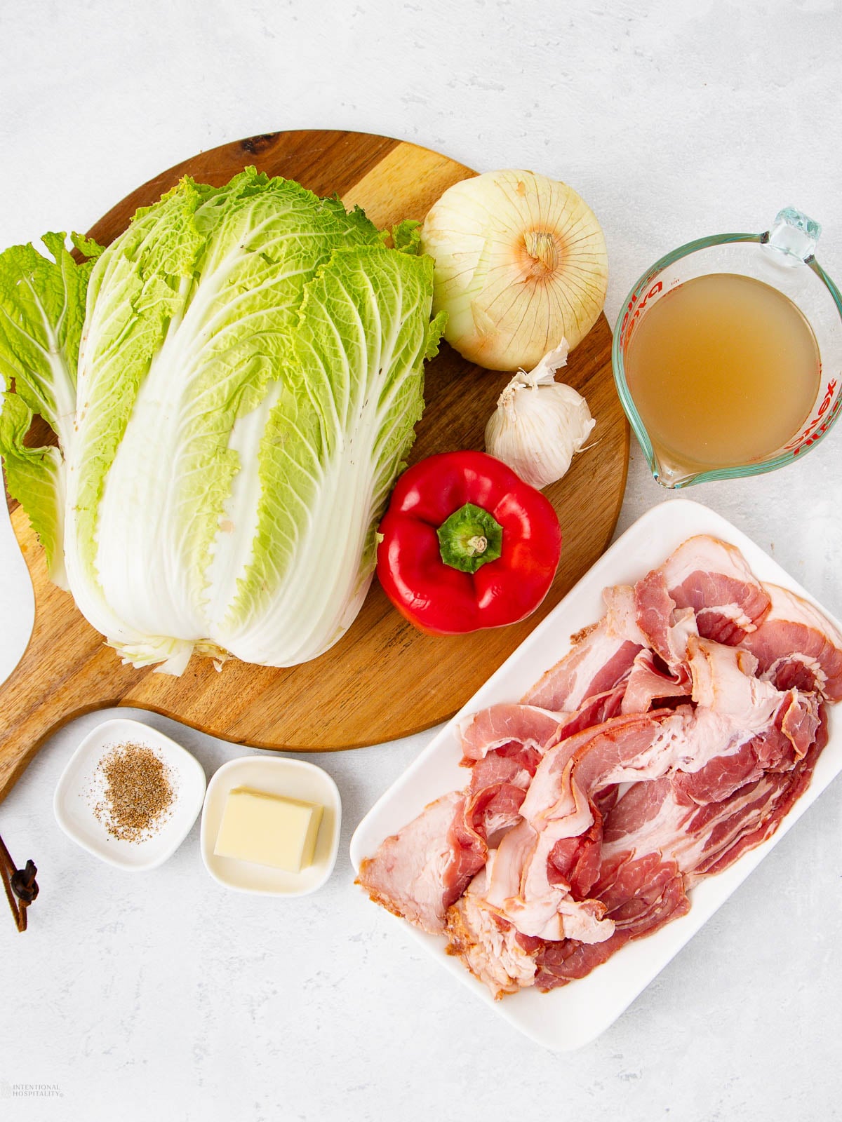 A wooden board holds Napa cabbage, a red bell pepper, an onion, and garlic. Next to it are a measuring cup of broth, a plate of bacon slices, a small bowl of butter, and a small bowl of seasoning.