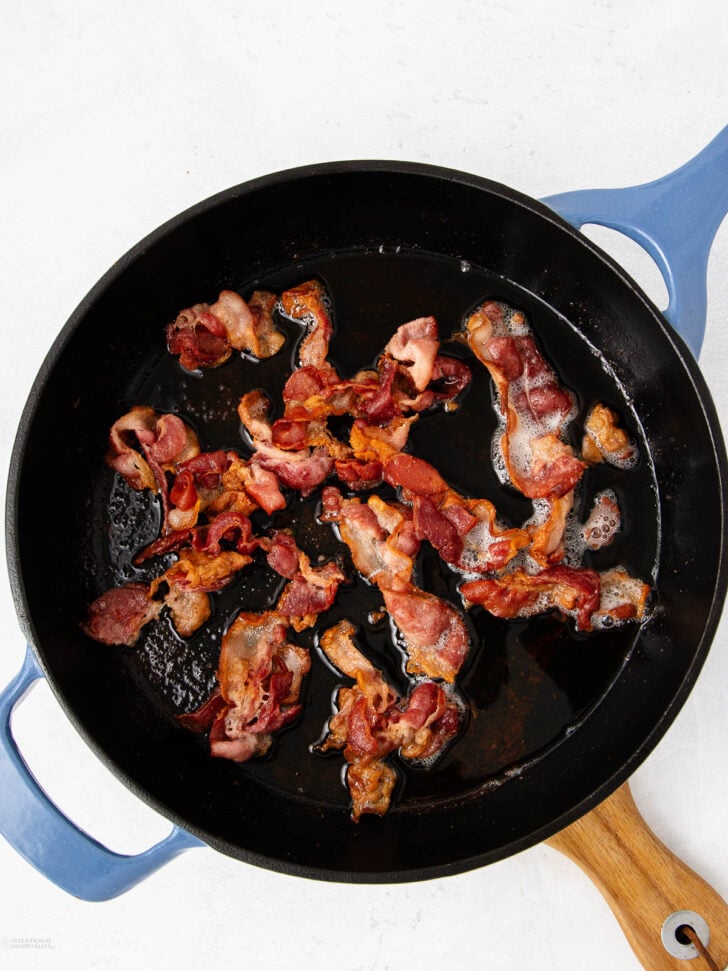 Crispy cooked bacon strips in a black skillet with a wooden handle, resting on a white surface.