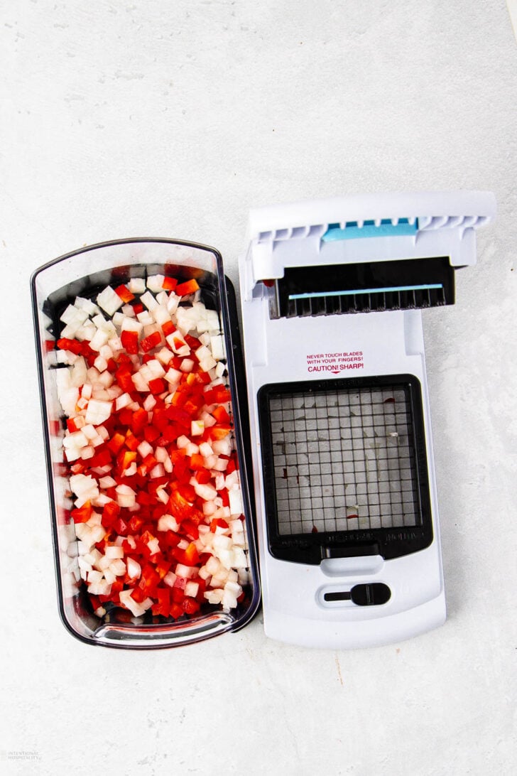 A vegetable chopper with a container filled with diced red and white vegetables, positioned on a light gray surface. The chopper's lid is open, revealing a grid of sharp blades.
